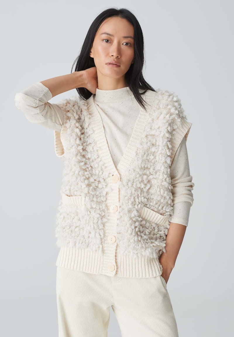 Cremfarbener ärmelloser Cardigan aus strukturiertem, flauschigem Material, mit Knopfverschluss und zwei vorderen Taschen. Getragen über einem eng anliegenden hellen Rollkragenpullover.