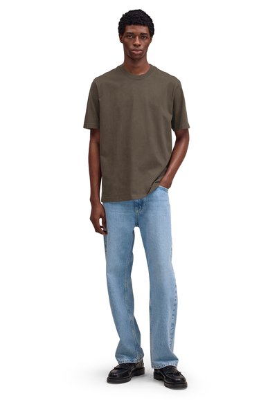 Młody mężczyzna stojący z ręką w kieszeni, ubrany w ciemnooliwkową koszulkę, jasnoniebieskie dżinsy i czarne buty na białym tle.