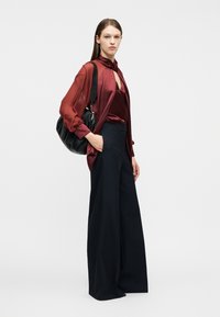 Γυναίκα που φοράει διάφανη μπορντό μπλούζα και φαρδιά μαύρη παντελόνα, κρατώντας μεγάλη μαύρη τσάντα ώμου, στέκεται μπροστά σε λευκό φόντο.