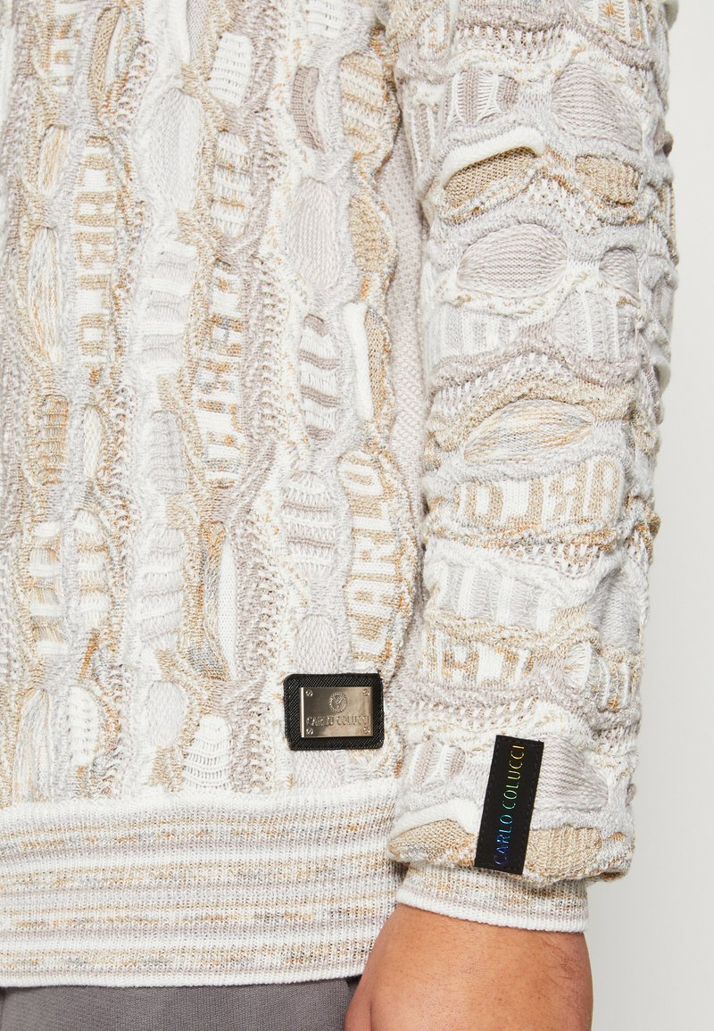 Trui in lichtbeige met een gewoven patroon, voorzien van horizontale en verticale kabels. Bevat een metalen logo label en een geweven etiket.