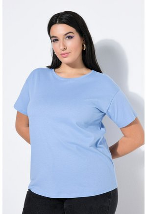 Woman with long dark hair wearing a light blue short-sleeve top and black pants, standing with hands behind back against white wall.