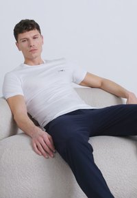 Νεαρός άνδρας που φοράει λευκό κοντομάνικο μπλουζάκι και σκουρόχρωμο παντελόνι, ξαπλωμένος σε έναν ανοιχτόχρωμο υφασμάτινο καναπέ με απλό φόντο.