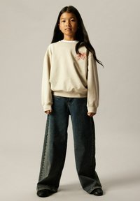 Ung jente med langt mørkt hår iført en oversized beige genser med rosa sløyfedekorasjon og vide mørke denimbukser som står.