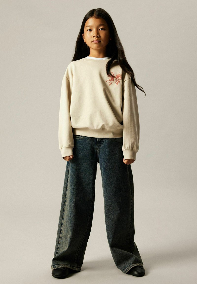 Ung jente med langt mørkt hår iført en oversized beige genser med rosa sløyfedekorasjon og vide mørke denimbukser som står.