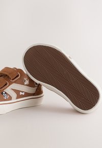 Chaussures marron pour tout-petit avec motifs de chien et d’os, attaches velcro et semelles en caoutchouc blanc, une chaussure montrant une semelle marron à motif zigzag.