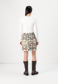 Femme aux longs cheveux foncés portant un haut blanc à manches longues, une jupe à motifs et des bottes noires montantes jusqu'aux genoux, vue de dos sur un fond blanc uni.