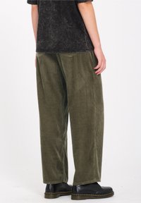 Osoba ubrana w oliwkowe, szerokie spodnie z korduru, czarny t-shirt w stylu acid-wash i czarne skórzane buty z żółtym przeszyciem, stojąca na białym tle.