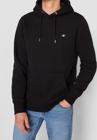 Sudadera negra de pullover hecha de tela suave, con un bolsillo frontal, capucha con cordón y pequeño detalle de logo en el pecho. Se muestran jeans azules.