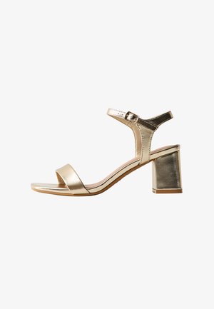 Sandalo aperto in metallo oro con cinturino alla caviglia, chiusura con fibbia e tacco largo su sfondo bianco.