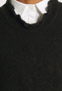 Gros plan d’un col de chemise blanche boutonné porté sous un pull noir à texture duveteuse.