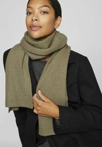 Olivengrøn rippet tørklæde lavet af blødt strikket stof, løs snoet om halsen; parret med en sort frakke og en grå top under.