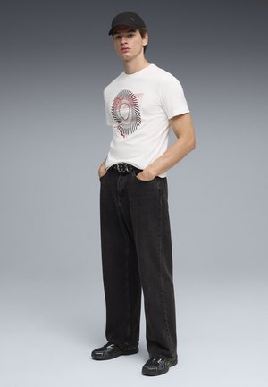Młody mężczyzna w białej koszulce z nadrukiem, czarnych szerokich dżinsach z paskiem z ćwiekami, czarnych trampkach i czarnej czapce, stojący z rękami w kieszeniach.