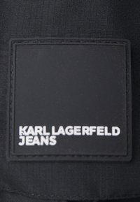Černá textilní etiketa přišité na tmavém textilu s bílým vyraženým textem "KARL LAGERFELD JEANS" psaným velkými písmeny.