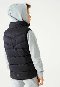 Melns polsterēts veste ar augstu apkakli un chevron šuvēm, uzvilkta virs pelēka kapuces krekla. Vestē ir tumšs uzraksts "BAKER" apakšmalā.