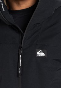 Czarna kurtka na zamek z kapturem, z fakturowaną powierzchnią, wyraźną naszywką z logo oraz kontrastowym zamkiem błyskawicznym.