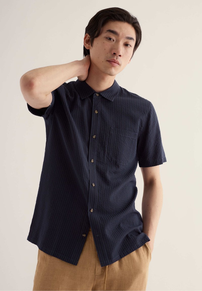 Homme portant une chemise bleu marine à manches courtes boutonnée et un pantalon beige, avec une main derrière la nuque, debout devant un fond clair uni.