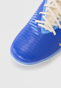 Scarpa sportiva blu con superficie testurizzata, caratterizzata da accenti blu chiaro e bianchi e lacci beige. Nota logo Nike in giallo.