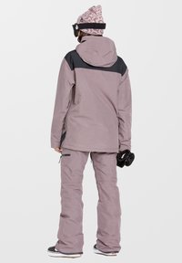 Chaqueta y pantalones de esquí en suave morado con acentos en negro, material impermeable, capucha ajustable, corte relajado y bolsillos con cremallera.