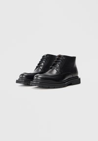 Botas de cuero negras hasta el tobillo con suelas de goma gruesas y cordones, presentadas sobre un fondo claro liso.
