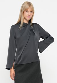 Blusa satin grigia scura con scollatura drappeggiata, maniche lunghe e fessure laterali. Texture liscia e design aderente, abbinata a una gonna grigia.