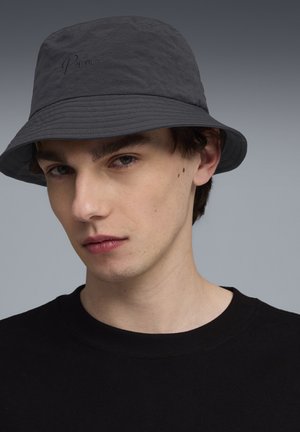 Jonge man met een donkere bucket hat met geborduurd logo en een zwart shirt met ronde hals, kijkt met een neutrale expressie recht voor zich uit.