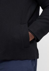 Czarna kurtka z mieszanki wełny o gładkiej fakturze, wyposażona w boczną kieszeń i luźny krój. Mankiety wykończone kontrastowym materiałem.