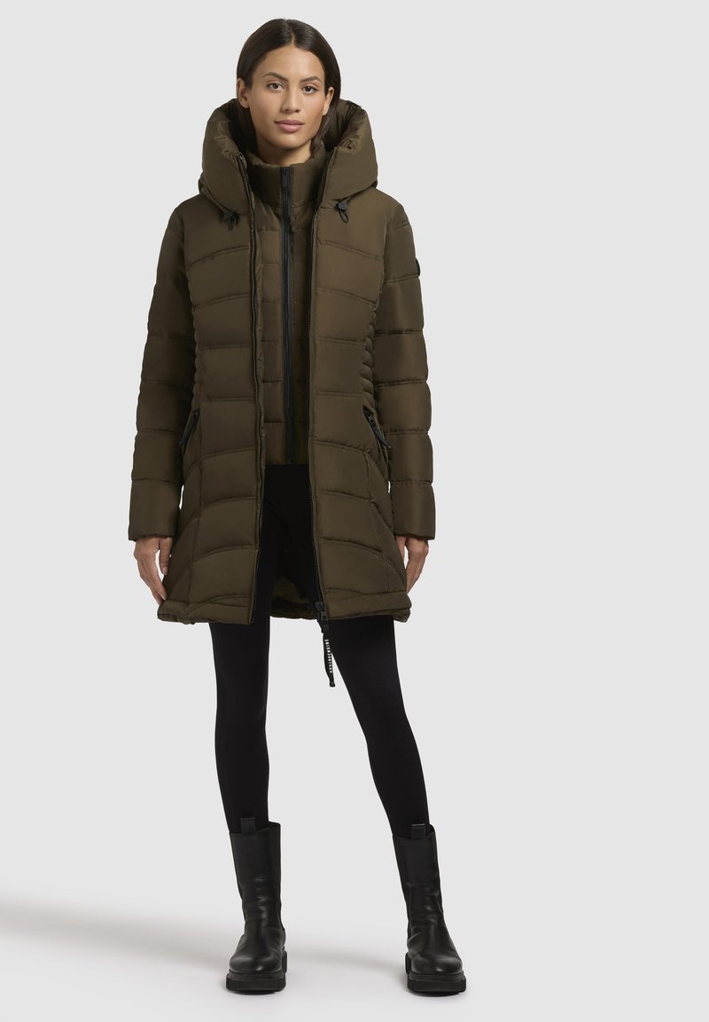 Olivgrüner Puffer-Mantel mit hohem Kragen, Reißverschlussfront und seitlichen Taschen. Strukturiertes Steppmuster und verstellbare Kapuze, kombiniert mit schwarzen Leggings und Stiefeln.