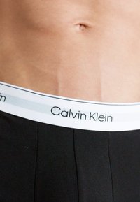 Crne pamukaste gaće s bijelim pojasom na kojem se nalazi logo "Calvin Klein". Glatka tekstura, prilagođen dizajn.