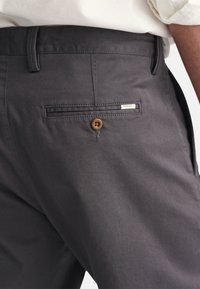 Gros plan sur la poche arrière d'un pantalon gris avec bouton et petite étiquette GANT, porté avec une chemise de couleur claire.
