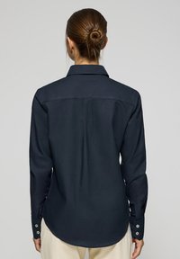 Camicia a maniche lunghe blu navy con colletto a punta, piega posteriore e polsini con bottoni, caratterizzata da una texture di tessuto liscia e orlo curvo.
