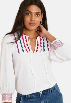 Vrouw met lang donker haar, gekleed in een witte blouse met kleurrijke, geschulpte borduursels op de borst en gerimpelde manchetten, gecombineerd met een blauwe spijkerbroek.