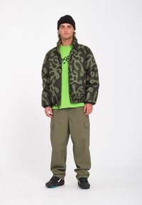 Gevoerde jas in donkergroen met een spiraalpatroon, gedragen over een felgroene t-shirt. Cargo-broek in khaki en zwarte schoenen maken de look compleet.