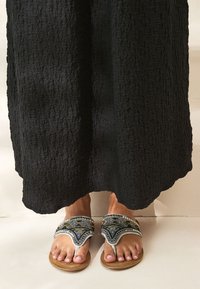 Sandales plates avec une tige ornée de perles argentées et bleues, présentant un design ajouré distinctif. La semelle est en matériau beige clair.