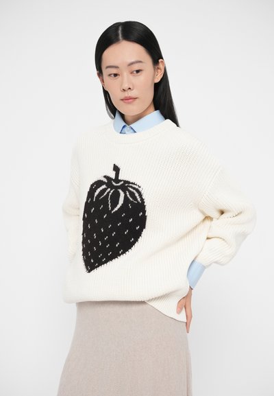 Camisola de malha creme com um grande desenho de morango preto. Possui decote redondo e corte largo. Usada sobre uma camisa azul clara com colarinho.