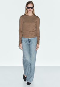 Brown long-sleeve knit top, light blue wide-leg jeans, and black high-heeled shoes. Model wearing sleek black sunglasses.