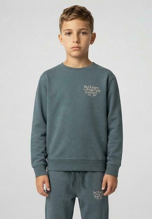 Jongen met kort lichtbruin haar, die een bijpassende blauwe trui en broek draagt met witte tekst "Be Honest and Fear Only Yourself."