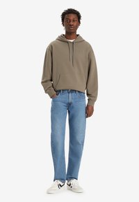 Sudadera caqui con cordón, bolsillo frontal, combinada con jeans rectos de color azul claro y zapatillas blancas con detalles en negro.