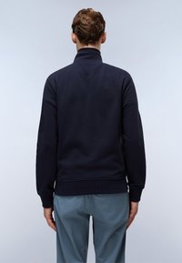 Felpa blu navy con colletto alto, polsini e orlo a costine, realizzata in tessuto morbido. Indossata con pantaloni azzurri chiari, leggermente lucidi.