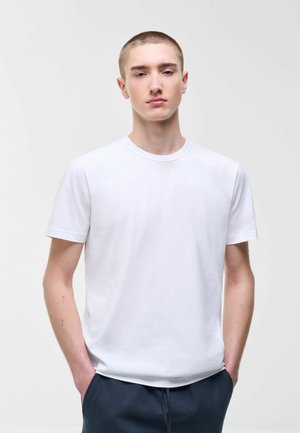 Giovane uomo con capelli corti che indossa una maglietta bianca semplice e pantaloni scuri, in piedi con le mani in tasca contro uno sfondo chiaro.