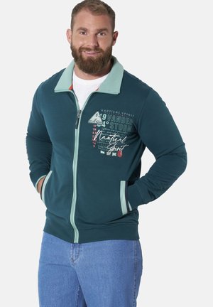 Man with beard wearing teal zip-up jacket with nautical text and graphics and blue jeans, hands in pockets, smiling against white background.
