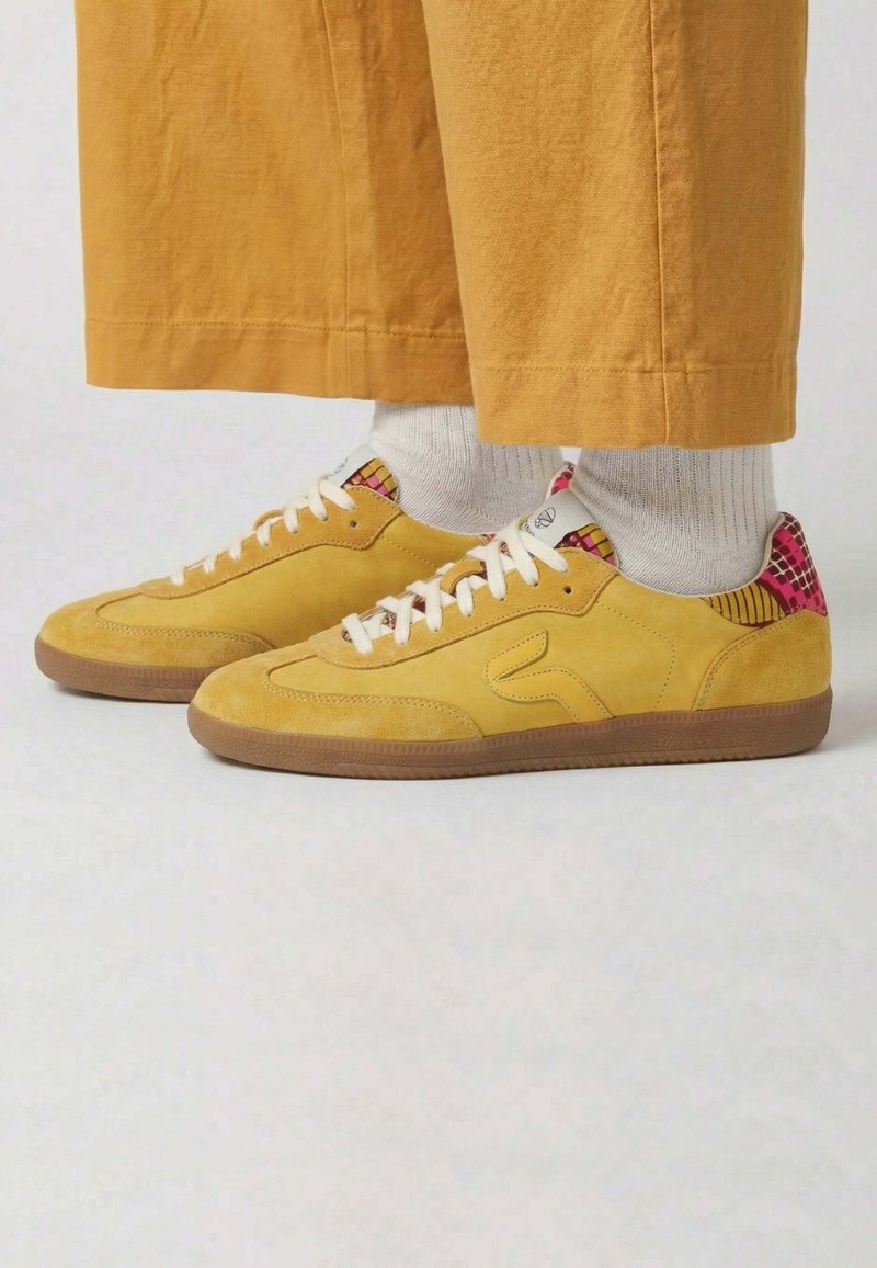Baskets en daim jaune avec des lacets blancs et un talon à motif coloré, portées avec des chaussettes blanches et un pantalon large couleur moutarde.