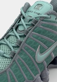 Zapatilla deportiva de Nike en tonos verde azulado y gris, con material de malla, diseño texturizado, sistema de cordones destacado y una estructura de suela distintiva.
