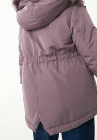 Veste rembourrée violette avec capuche, bordure en fourrure et taille élastique. Elle présente une texture douce, un ourlet arrondi et des détails de fente sur les côtés.