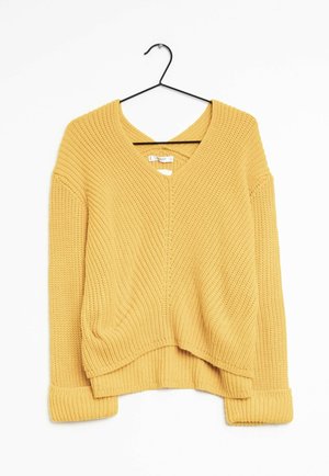 Pull jaune en maille à col en V avec manches longues et poignets retournés, suspendu à un cintre en métal noir sur fond blanc.