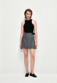Schwarzes geripptes Tanktop und grauer plissierter Minirock mit einem schwarzen Gürtel. Das Model trägt schwarze Loafer. Einfache, klare Linien und klassische Farben.
