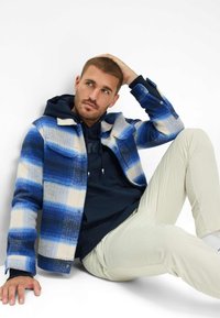 Giacca a scacchi blu e bianca con trama a spina di pesce, indossata sopra una felpa blu navy, abbinata a pantaloni chiari in velluto a coste.