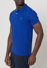 Kunga blå polo skjorta i mjuk bomull, med en grön logotyp, knäppning fram och kontrasterande krage. Korta ärmar och normal passform.