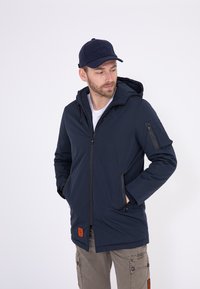 Marineblaue wasserdichte Jacke mit Kapuze, Reißverschluss vorne und Seitentaschen. Verfügt über eine aufgesetzte Tasche am Ärmel und ein strukturiertes Logo-Patch.