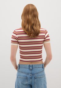 Mujer con cabello rojo hasta los hombros que lleva una camiseta corta de manga corta a rayas y jeans azul de cintura alta, mirando hacia una pared blanca lisa.