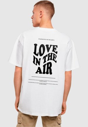 T-shirt en coton blanc avec un texte noir au dos qui proclame "AMOUR DANS L'AIR" dans une police ondulée. Coupe décontractée et manches courtes.